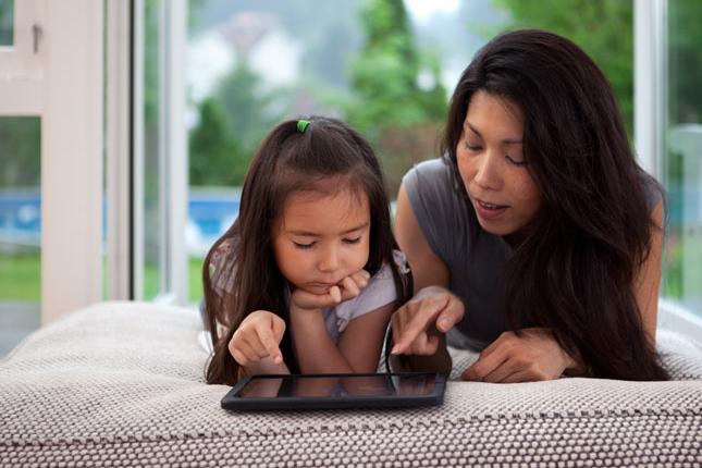 A mother and her young daughter lay across a sofa while looking at a tablet.