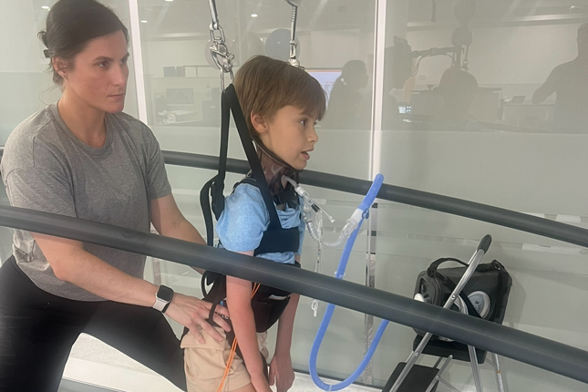 Un niño participa de una sesión de TSS sobre una caminadora, mientras su fisioterapeuta está detrás.