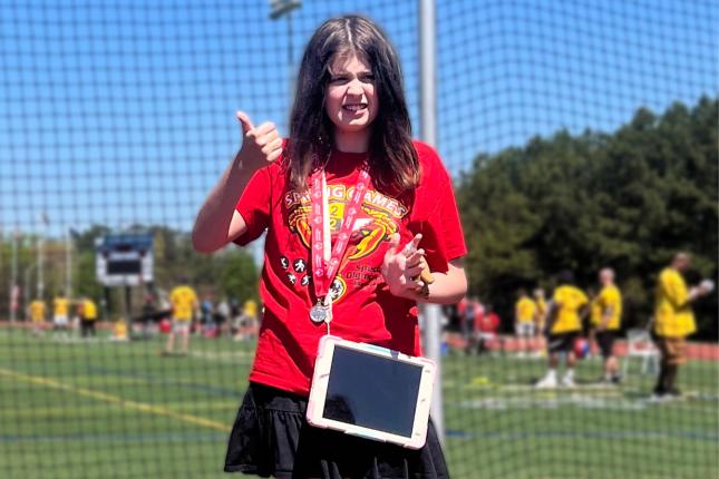 A girl wearing a augmentative and alternative communication device across her shirt stands up for a posed photo. She is participating in a Special Olympics event, and is also wearing a medal. 