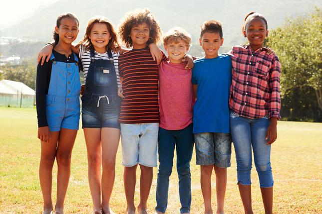 Six pre-teen friends stand shoulder to shoulder while smiling in a park.