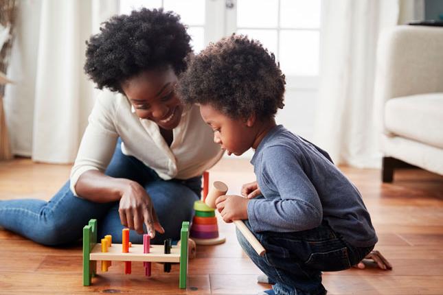 A mother smiles while sitting on the floor with her toddler son and playing with a toy.