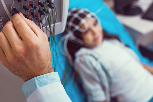 Close up of a laboratory worker's hand setting up an electroencephalography machine before analyzing brain of a little girl lying on an examination couch in the background.