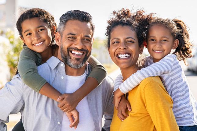 A father, mother and two daughters smile in an outdoor photo. Each parent has one of the daughters on their back.