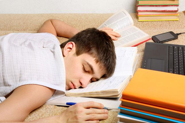 A tired teenage boy rests his head on an open notebook while studying for an exam.