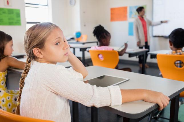 A little girl gazes off to the side as she sits in a classroom. She is looking away from her teacher, who is writing on a whiteboard as the other students watch.