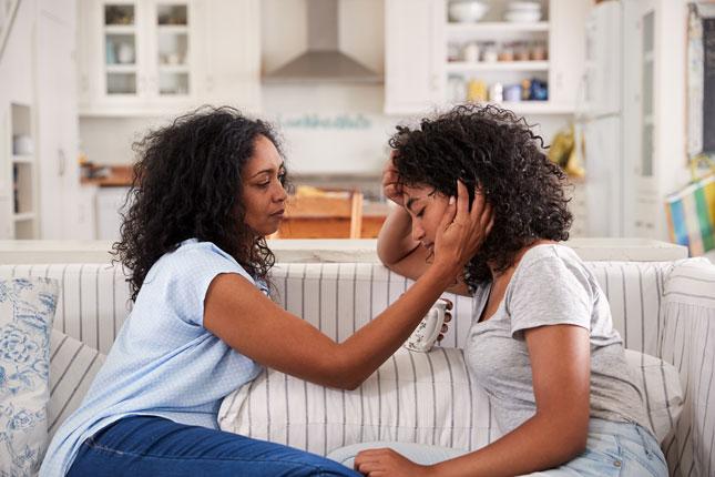 A mother comforts her upset teenage daughter as they both sit on a sofa.