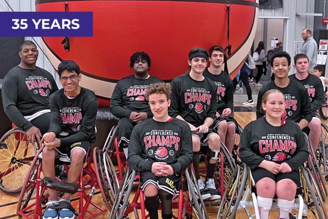 Nine teens pose for the camera. Each sits in a wheelchair, and they appear to be on a basketball court. Each teen wears a black T-shirt that says “Conference Champs” on it.