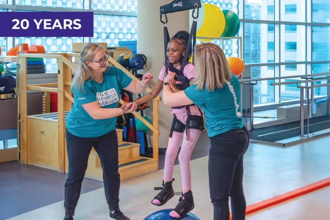 A smiling young girl is suspended in a harness from a weight-supporting device. On her feet are black braces that support her feet and ankles. Helping her are two women, both wearing dark pants and teal T-shirts that say “Team Kennedy Krieger.” In the background are colorful therapy balls and other physical therapy equipment.