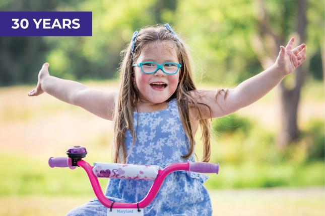 A smiling young girl wearing aqua-framed glasses sits on a purple and pink bike outside, her arms stretched widely. The bike is stationary.
