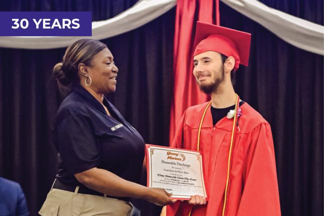 A woman in a short-sleeved navy blouse and khaki pants and wearing her hair in a bun hands a certificate to a young man wearing a red graduation cap and gown. They appear to be standing on an auditorium stage, and both are smiling at each other.