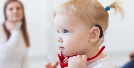 A profile of a girl toddler wearing a hearing aid. She is looking straight ahead. The background of the photo is blurred, and includes a woman who is looking at the child while raising her right hand towards her neck.