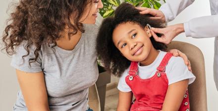 A little girl with her parent while a hearing aid is installed in her left ear by an audiologist.