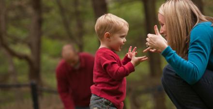 A young boy and his mother communicate through American sign language. They are outside, with the boy's father standing in the background.
