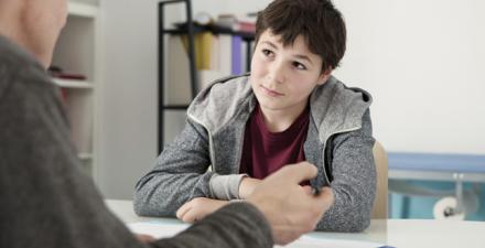 A boy sits across the desk from a man who is speaking. The man's back is turned to the camera, and he is holding a pen, with a clipboard in front of him on the desk.