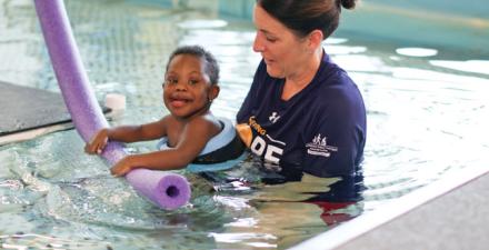 A child with floatation devices swimming in a pool with a helper. 