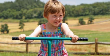 A young girl holds the handlebars of a green tricycle. Behind her is a fence, with a hilly farm field on the other side.