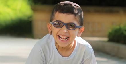 A young boy laughs and smiles while playing outside. He wears glasses and a kippa, and has a birthmark on his right ear and on the right side of his neck.