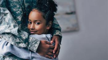 A young girl looks at the camera as she hugs an adult wearing a military uniform