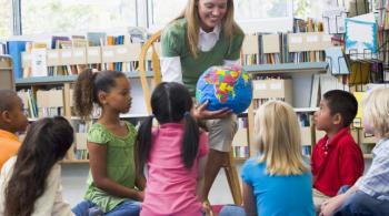 A photo taken in a classroom captures a teacher sitting in a chair and holding a globe and showing it to her students, sitting on the floor