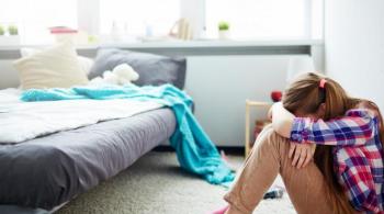 A photo of a girl sitting on the floor of her bedroom with her head down in her arms, in pain