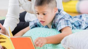 A child lays atop a pillow while reading a book, accompanied by an adult