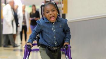 A photo of Journee in the hallway at Kennedy Krieger, smiling at the camera as she walks with the assistance of a walker