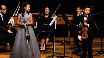 Musicians Maggie and Matthew Schneider stand on a stage holding violins, with an ensemble behind them.