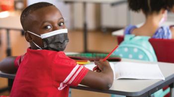 A young boy at his school desk looks over his shoulder at the camera while holding a pencil in his right hand and wearing a face mask