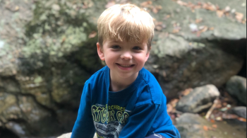 A photo of Hudson, a young Kennedy Krieger patient. Hudson has blonde hair and sits atop a rock.