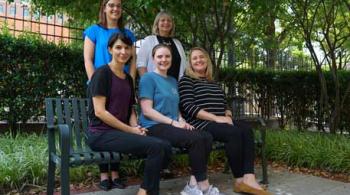 Four women pose outside, three sitting on a bench, and two standing behind the bench. All are smiling.