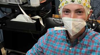 A young man sits in chair in a clinical room. He wears a face mask and a snug-fitting EEG cap fitted with dozens of electrodes. 