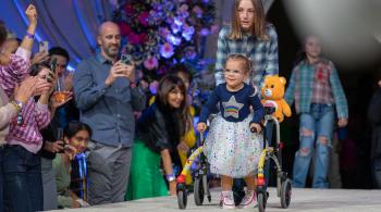 Penny, a child in colorful clothing, walks down a runway with assistance while lots of people in a crowd smile and watch.