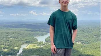 A young man smiles while standing atop a hill or summit. Behind him is a beautiful landscape, with trees, mountains and a river. 