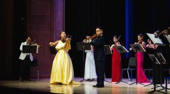 Siblings Maggie and Matthew, wearing a yellow dress and black suit respectively, stand on a stage playing violins with an ensemble in the background.