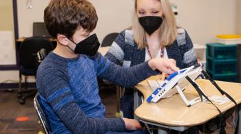 Jack sits at a student desk in a classroom with his teacher and uses a tablet-like communication device.