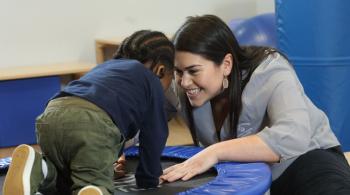 A child with autism plays on a small trampoline, while a woman at one end of the trampoline smiles at him. 