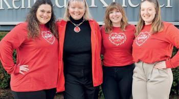 Four women stand outside in front of a sign that says “Kennedy Krieger.” Three of them are wearing red T-shirts that say “I love the holidays,” with the Kennedy Krieger Festival of Trees logo underneath.