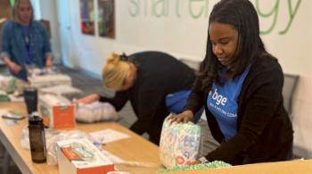 Two women work at a table assembling diapers into care packages. Each woman is wearing a blue T-shirt. One of the T-shirts can be seen to read “BGE, an Exelon Company.”