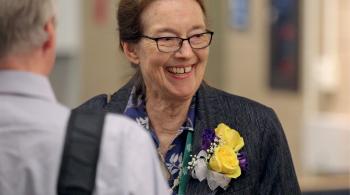 A woman smiles and looks off to the side. She is wearing a corsage of yellow roses and purple flowers, with a white bow. In the foreground is a man’s blurry back. They are standing in a room or hallway.