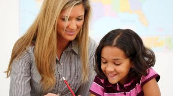 A woman teacher sits next to a young girl while she writes. 