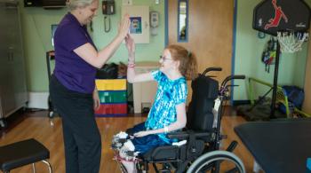 A teenaged girl in a blue and white shirt sits in her wheelchair and high fives her therapist, a woman wearing a purple shirt.