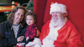 A father holds his toddler while kneeling next to Santa Claus at Festival of Trees.