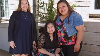 A teenaged girl in an elegant outfit sits in a wheelchair between two women. She leans toward the woman on the right, who leans toward the girl, as they are mother and daughter. All three are smiling.