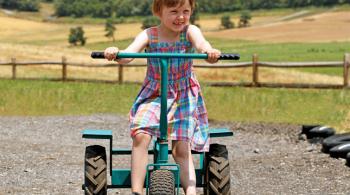 A smiling little girl with short blonde hair rides a tricycle in a rural setting.