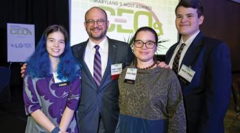Family photo of a teenaged girl and boy with their parents. Father and son are each wearing a suit and tie, and mother and daughter are each wearing a dress. Each family member is wearing a name badge. Behind them is a screen that says “The Daily Record—Maryland’s Most Admired CEOs.”