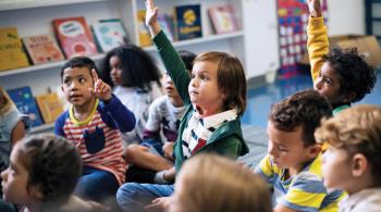 Three kids sitting on a floor in a classroom raise their hands. Several students sitting around them are not raising their hands.
