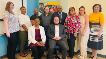 A group photo from a Faith Community meeting. A woman and man sit in the front with a group of four people standing behind them. The top of a green and yellow MCDD banner can be seen in the very back.
