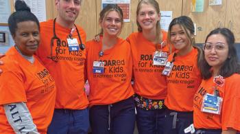 Six smiling people pose for the camera wearing bright orange shirts that say “I ROARed for Kids at Kennedy Krieger 2022.” Most of the people have badges on; some of the badges say “RN” on them. One of the people has a stethoscope around their neck.