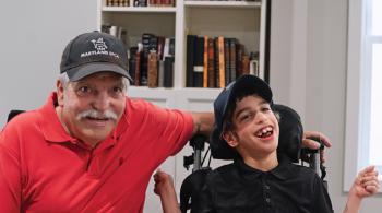 A man wearing a baseball hat that says “Maryland SPCA” on it sits beside a young boy who is also wearing a baseball cap. The boy is sitting in a wheelchair. The man’s left arm rests on the back of the wheelchair, behind the wheelchair’s headrest. Both the man and the boy are smiling. They are sitting indoors, in front of a bookshelf.