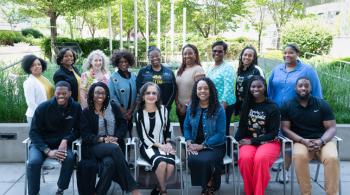 A group of 15 people pose in a garden-like setting. Six of them are sitting in chairs, while the other nine stand behind them. All are smiling, and most are people of color.